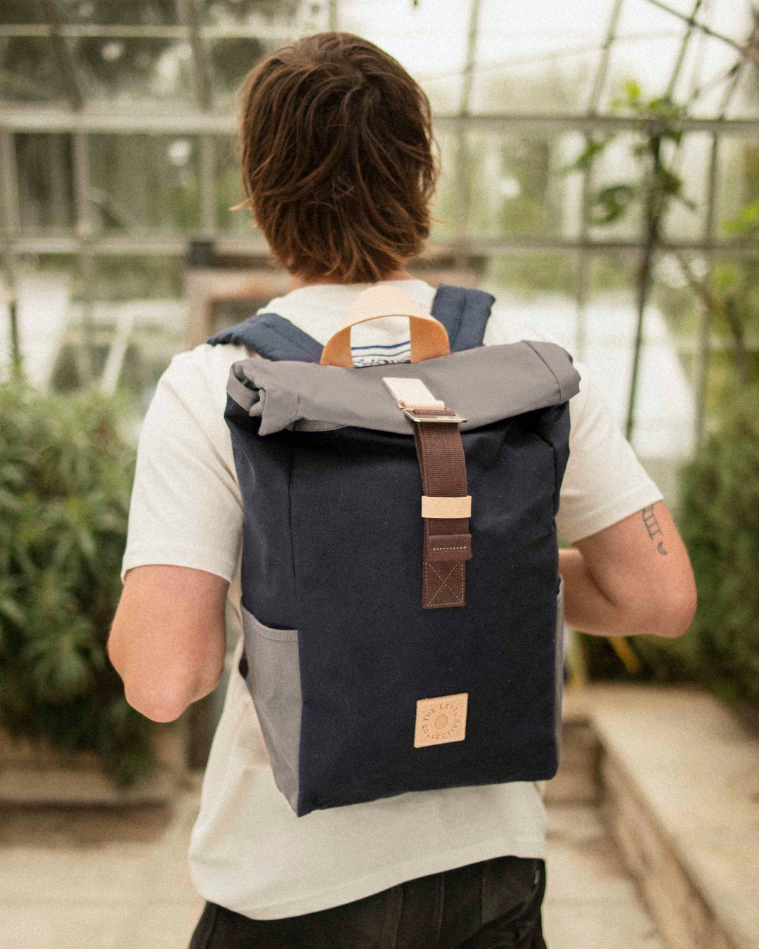 Man in a greenhouse wearing the Winnats Roll Top in Navy & Stone, navy waxed canvas with a stone roll top, stone side pockets and brown webbing
