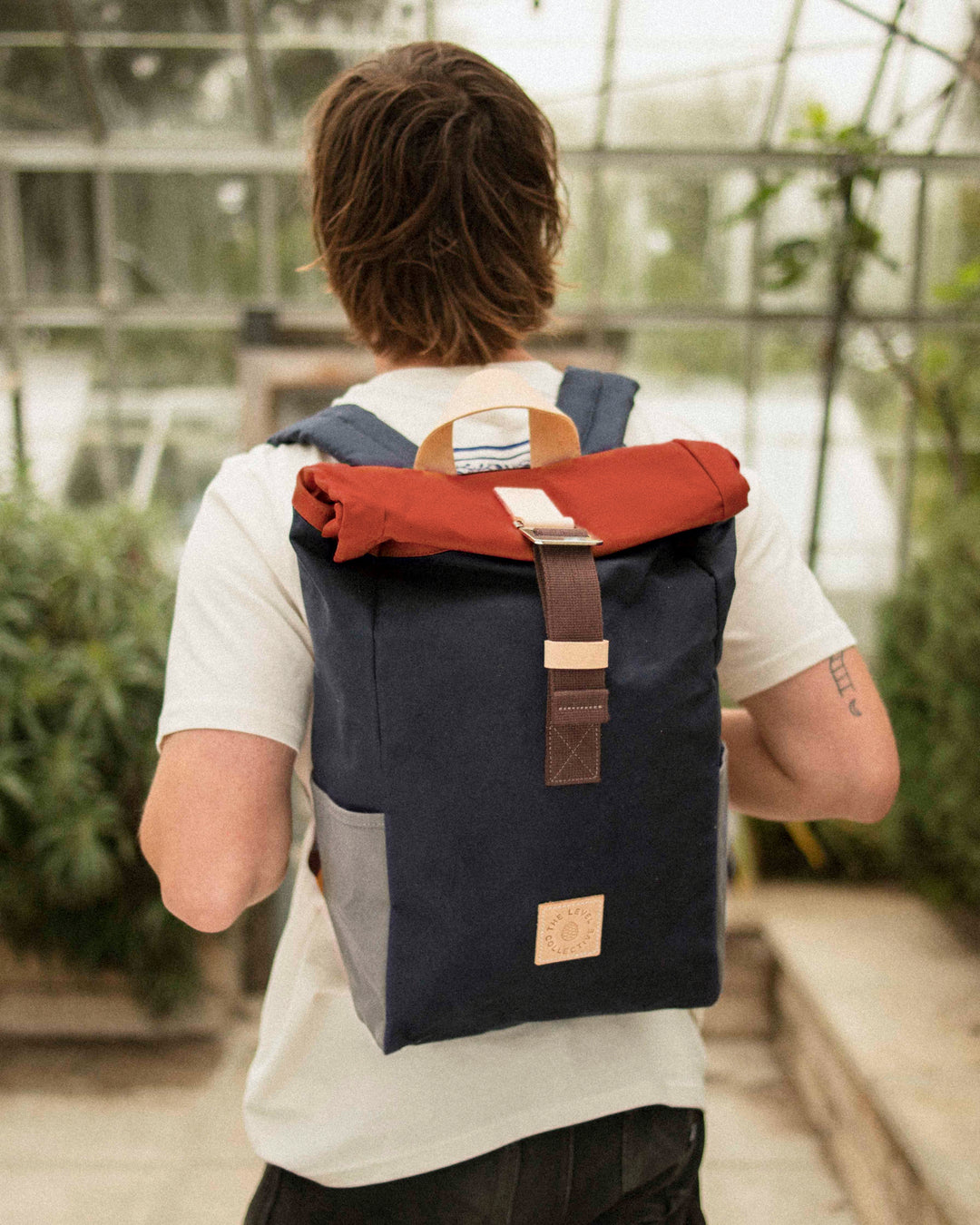 Man in a greenhouse wearing the Winnats Roll Top in Wild One Navy, navy waxed canvas with a rust roll top, stone side pockets and brown webbing