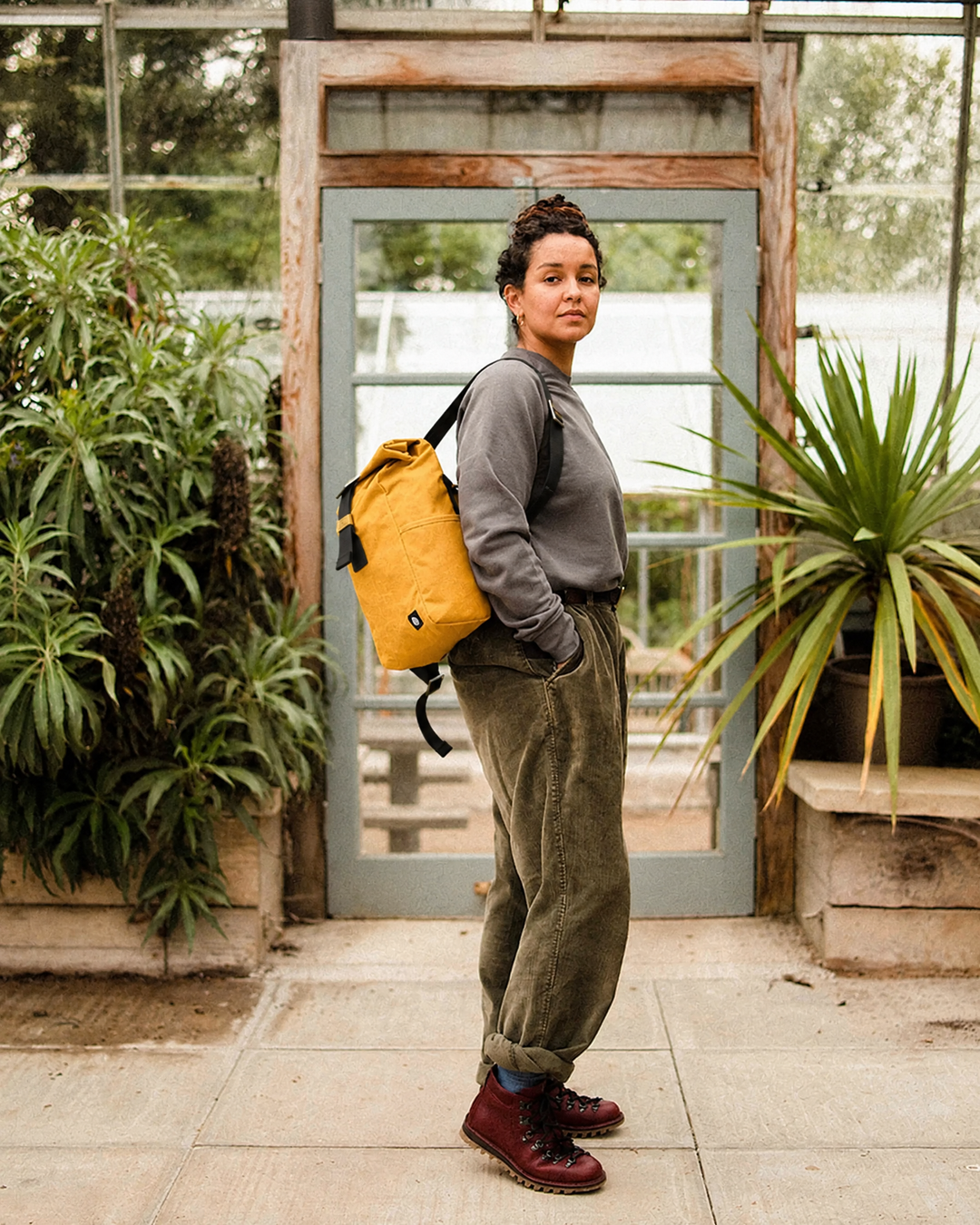 Roll Top Mini in Mustard worn on one shoulder at a greenhouse door